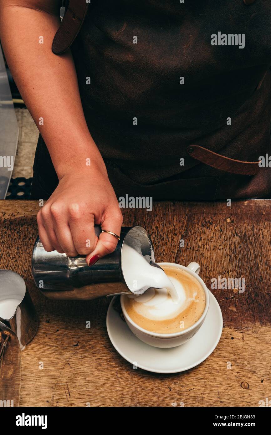 Barista making latte or cappuccino coffee, pouring steamed milk, making latte art. Cup of coffee on wooden table. Stock Photo
