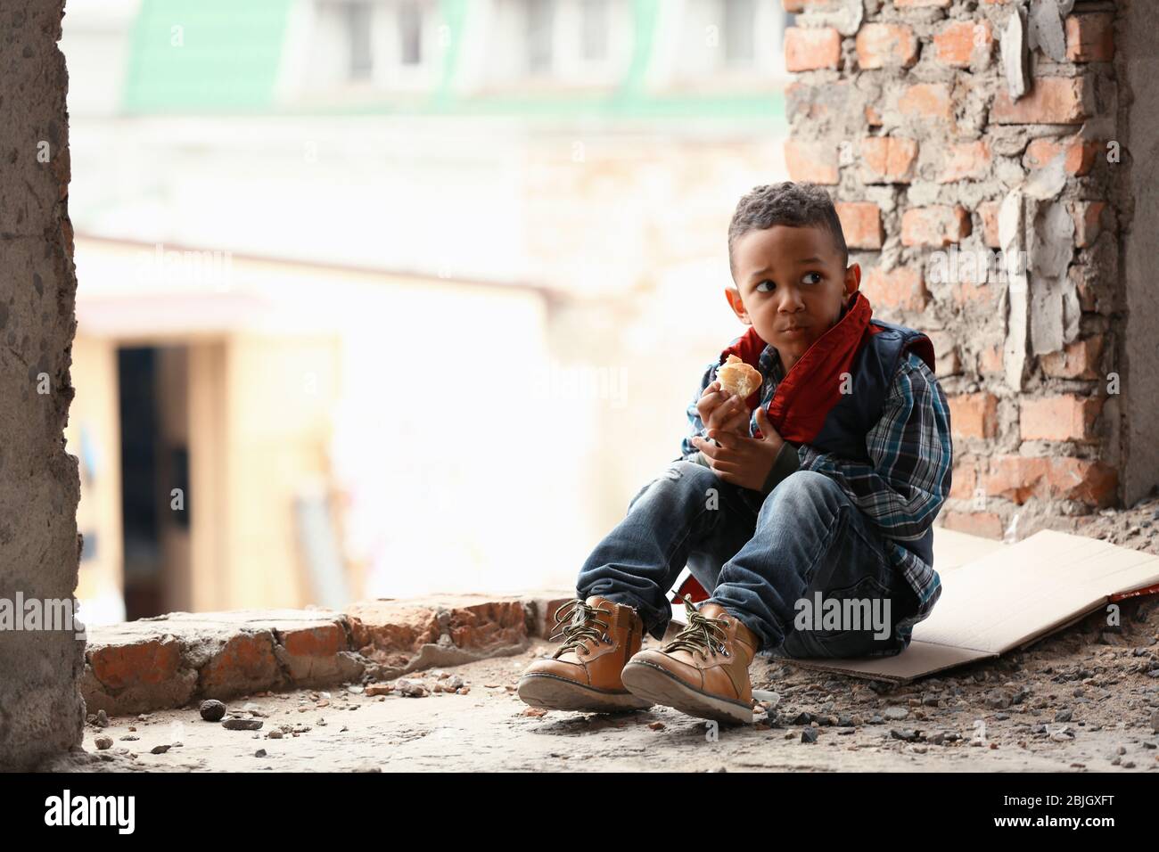 Cute little boy sitting on floor with piece of bread in abandoned building. Poverty concept Stock Photo