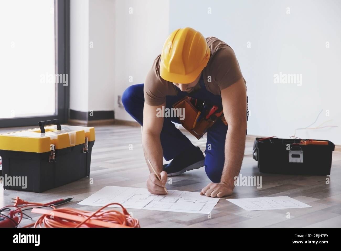 Young electrician checking drawings near toolboxes in light room Stock Photo
