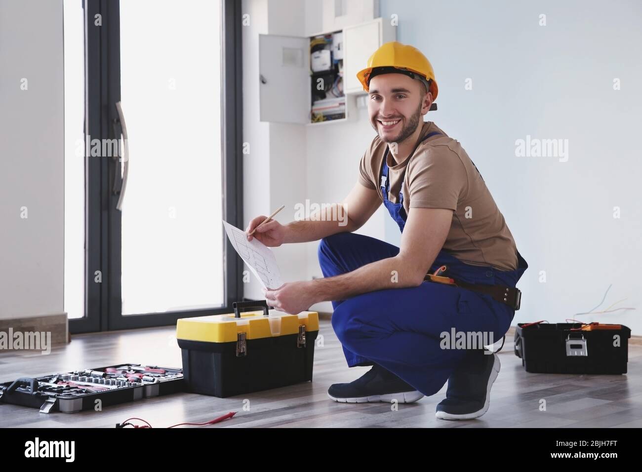 Young electrician checking drawings near toolboxes in light room Stock Photo