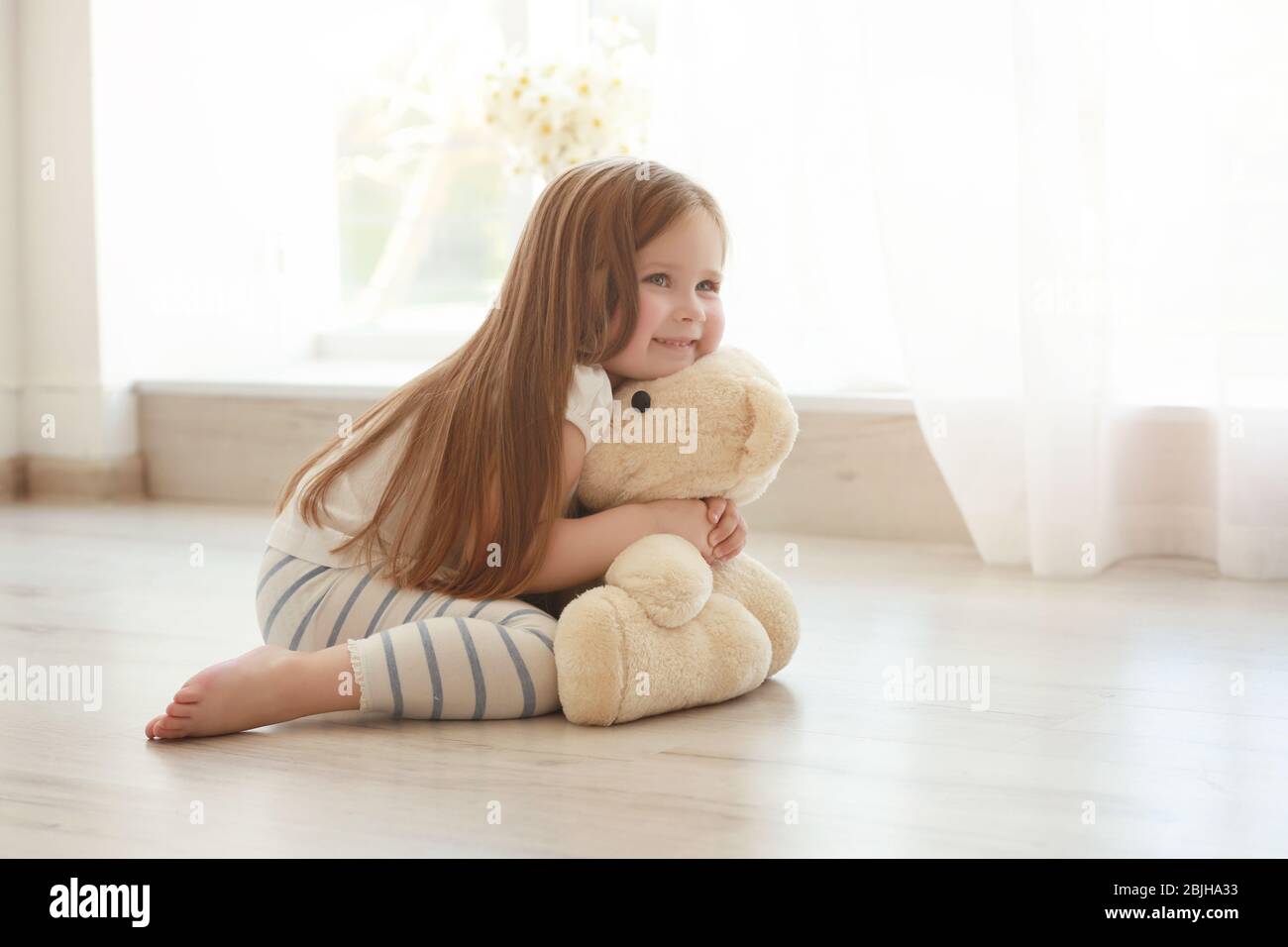 Cute little girl hugging teddy bear while sitting on floor at home ...