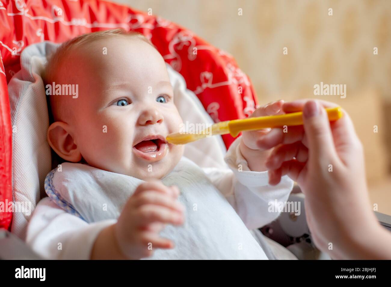 Mother feeding her happy baby son with spoon. Mother giving healthy food to her adorable child at home. Parent feeding baby boy in high chair. Close-u Stock Photo