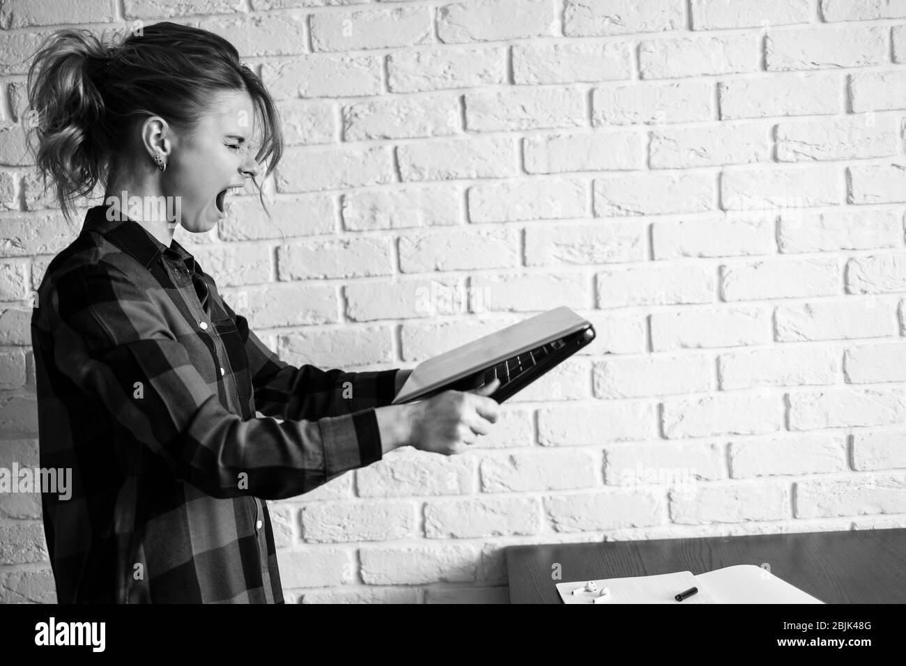 Black and white photo of female freelancer with raised laptop in hands standing agains white brick wall. Distant work Stock Photo