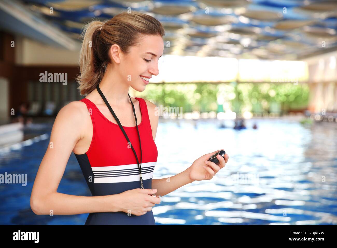 Beautiful young coach with stopwatch and whistle standing near swimming