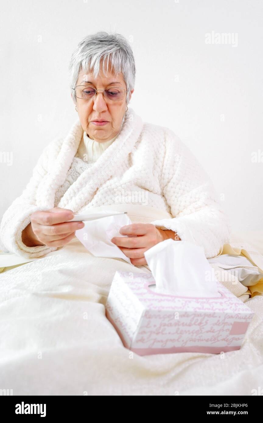 Coronavirus. Sick senior woman holding tissue and thermometer sit on bed, upset old mature woman caught cold got coronavirus flu influenza symptoms Stock Photo
