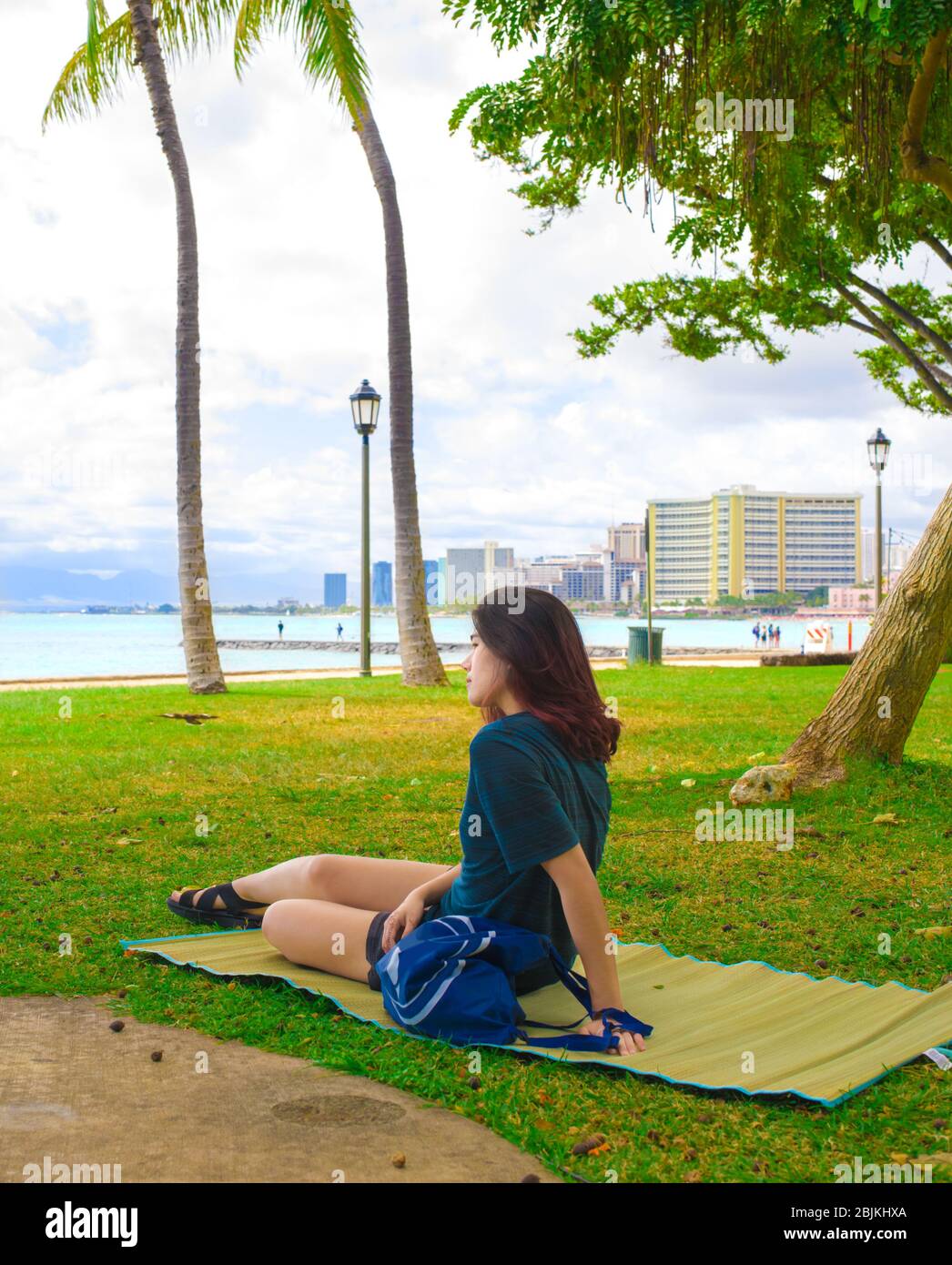 One biracial Asian teenage girl or young adult sitting alone on straw mat on grass lawn at empty Waikiki park looking out at ocean on cloudy overcast Stock Photo