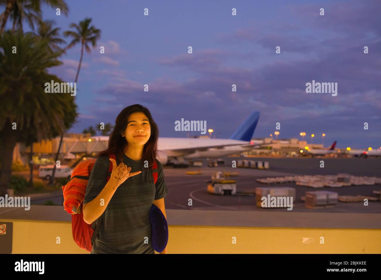 Teenage girl standing at Hawaiian airport making shaka sign before evening flgiht. Dark skies and airplanes in background Stock Photo