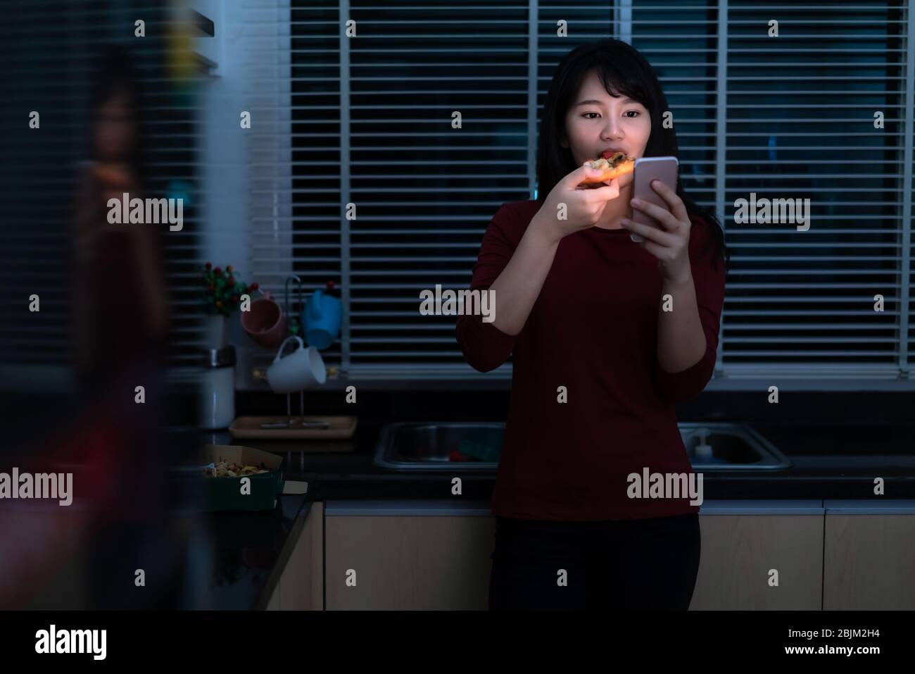 Asian woman virtual happy hour meeting and eating delivery pizza from the box online with friend or taking photo using mobile phone camera in kitchen Stock Photo