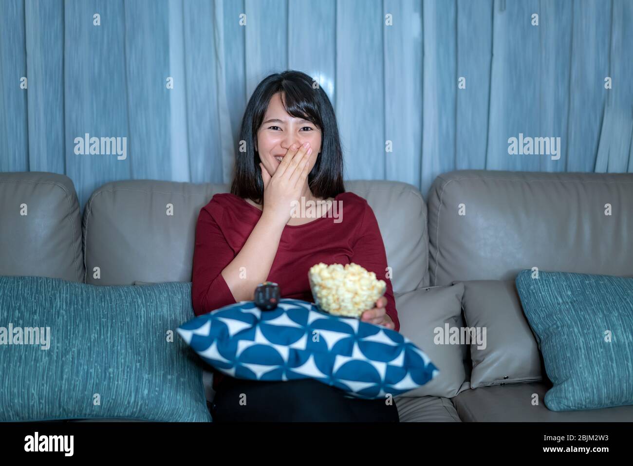 Young asian woman watching television suspense movie or news looking happy and funny and eating popcorn late night at home living room couch during ti Stock Photo
