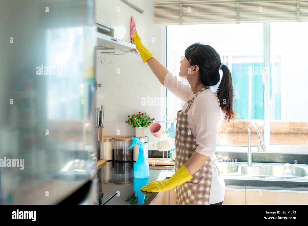 Asian woman wearing rubber protective gloves cleaning kitchen cupboards in her home during Staying at home using free time about their daily housekeep Stock Photo