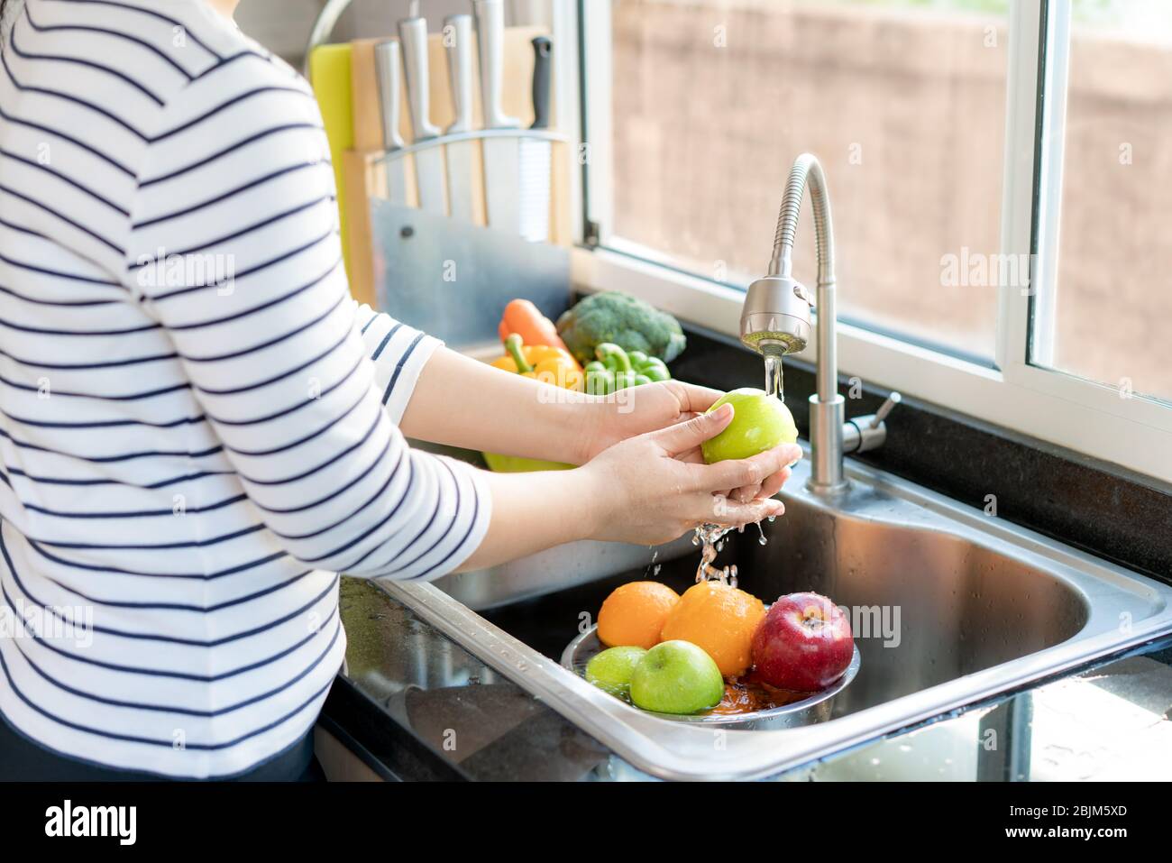 Asian healthy woman washing an apple and other fruit above kitchen sink and cleaning a fruit / vegetable with water to eliminate the chances of contam Stock Photo