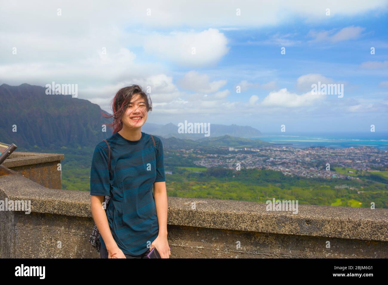 Biracial Asian Caucasian teen girl or young adult standing at Pali Lookout, Oahu, Hawaii on sunny, windy day with views of Kaneohe city and blue ocean Stock Photo