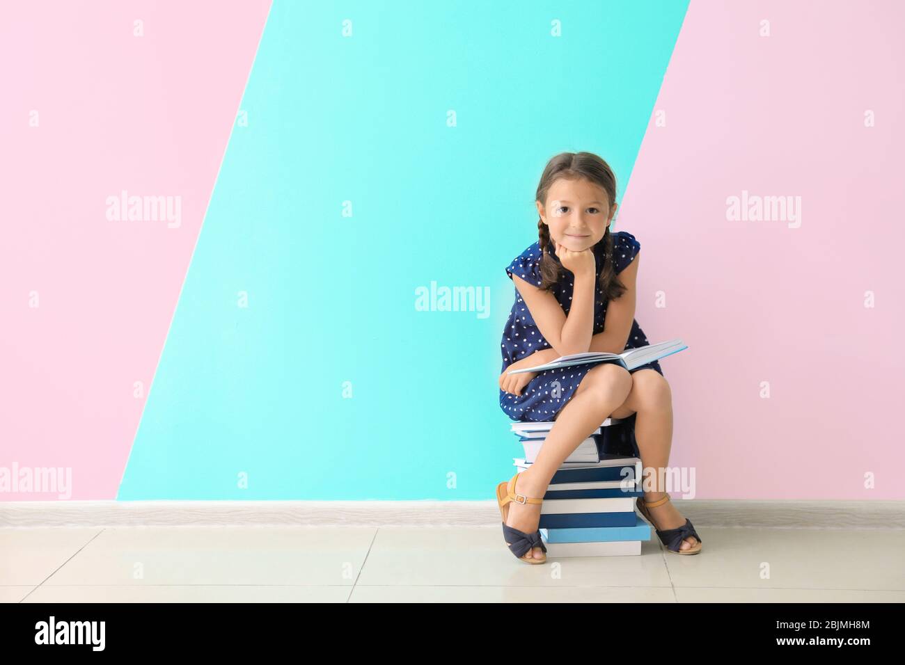 Cute little girl sitting on pile of books near color wall Stock Photo ...