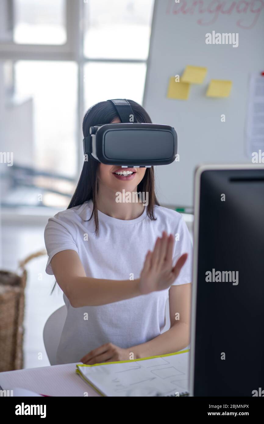 Dark-haired girl sitting at the table wearing vr headset and stretching her arm Stock Photo