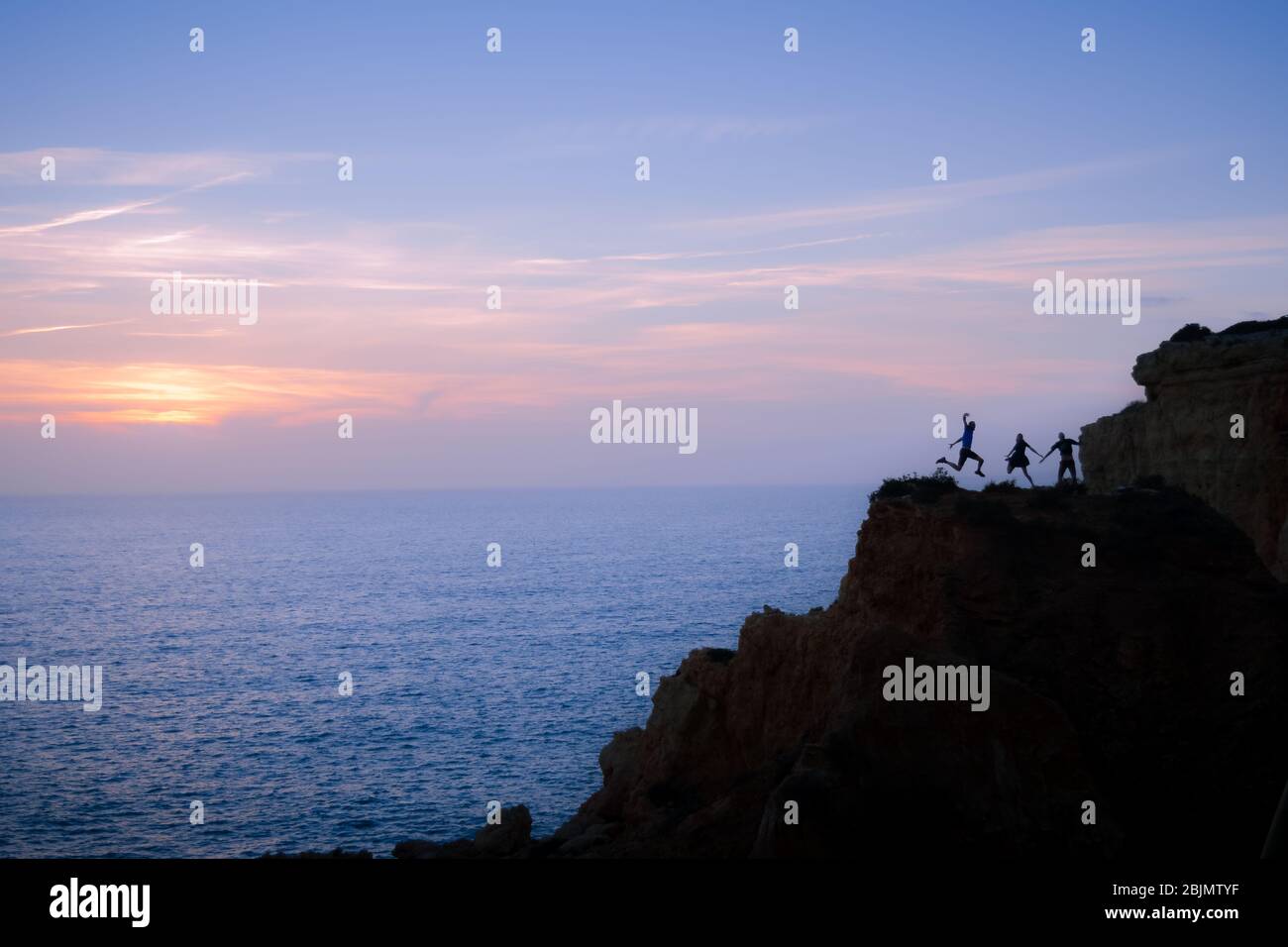 A group of 3 friends on vacation jumps at the same time for a picture taken on timer, making up a fun backlight image on top of the cliffs. Stock Photo