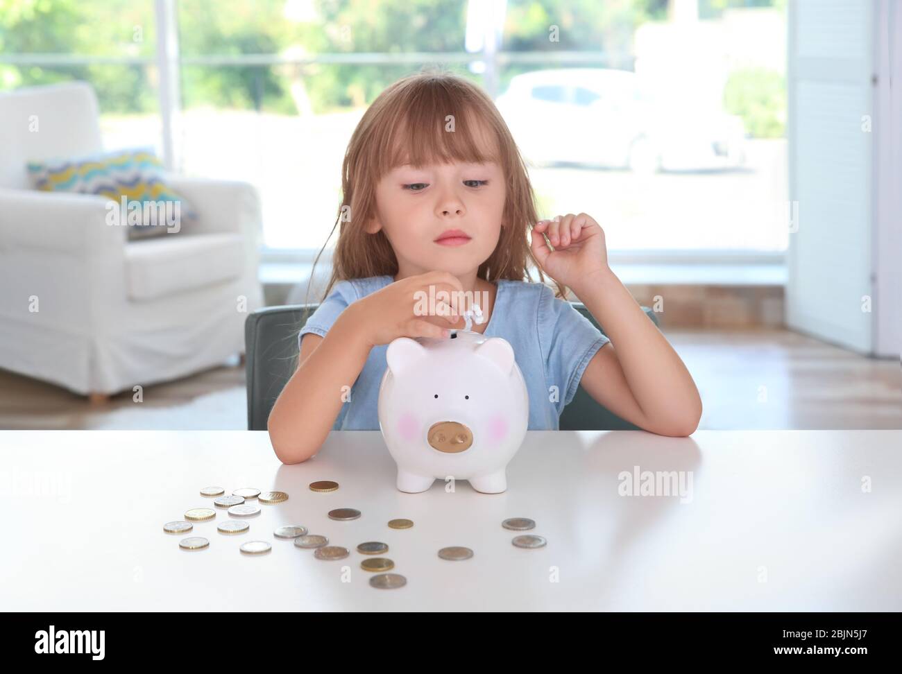 Cute little girl putting coin into piggy bank indoors Stock Photo - Alamy