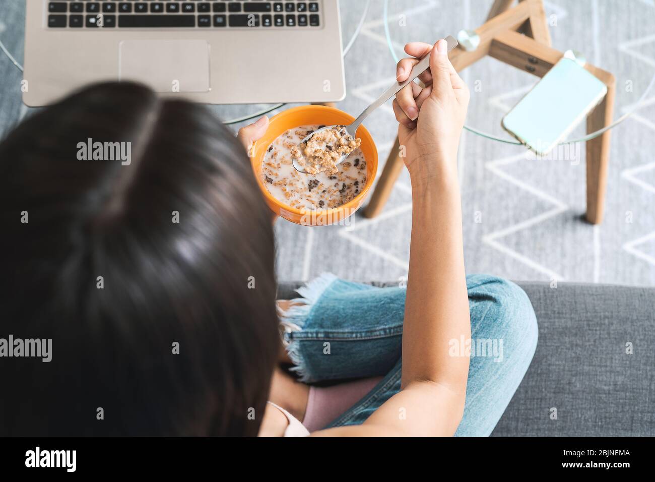 Top view female eating cereal cup with milk while watching on computer sitting on sofa - Health meal and living room entertainment concept Stock Photo