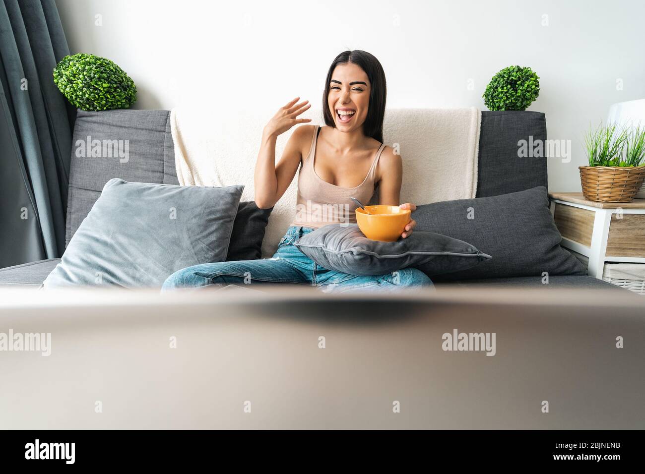 Happy woman eating cereal cup with milk while watching on computer laptop sitting on sofa - Health meal and living room entertainment concept Stock Photo