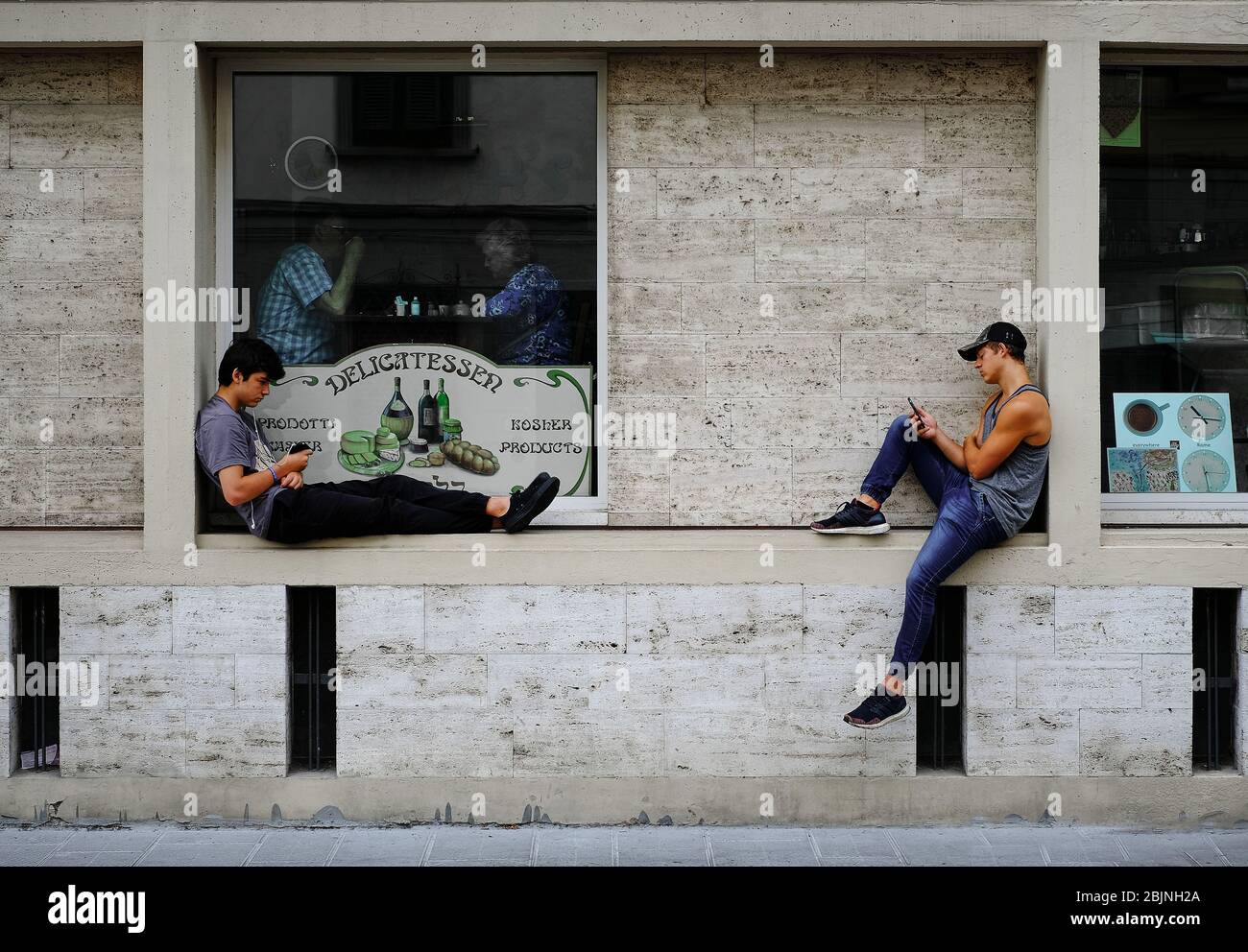 Two young men lounging on the wall of a restaurant below the window with people eating texting on their mobile phone in a city lifestyle and communica Stock Photo