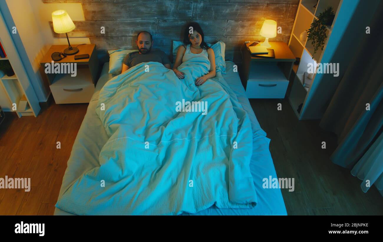 Young wife with her husband under the sheets telling him she wants a divorce. Blue moonlight in the room. Stock Photo