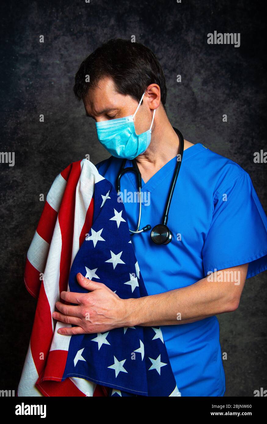 Sad/pensive male doctor in blue hospital scrubs with face mask and stethoscope, nursing the Stars & Stripes American flag close to his chest. Stock Photo