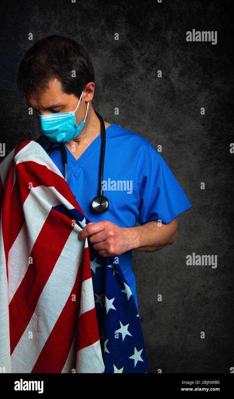 Sad/pensive male doctor in blue hospital scrubs with face mask and stethoscope, nursing the Stars & Stripes American flag close to his chest. Stock Photo