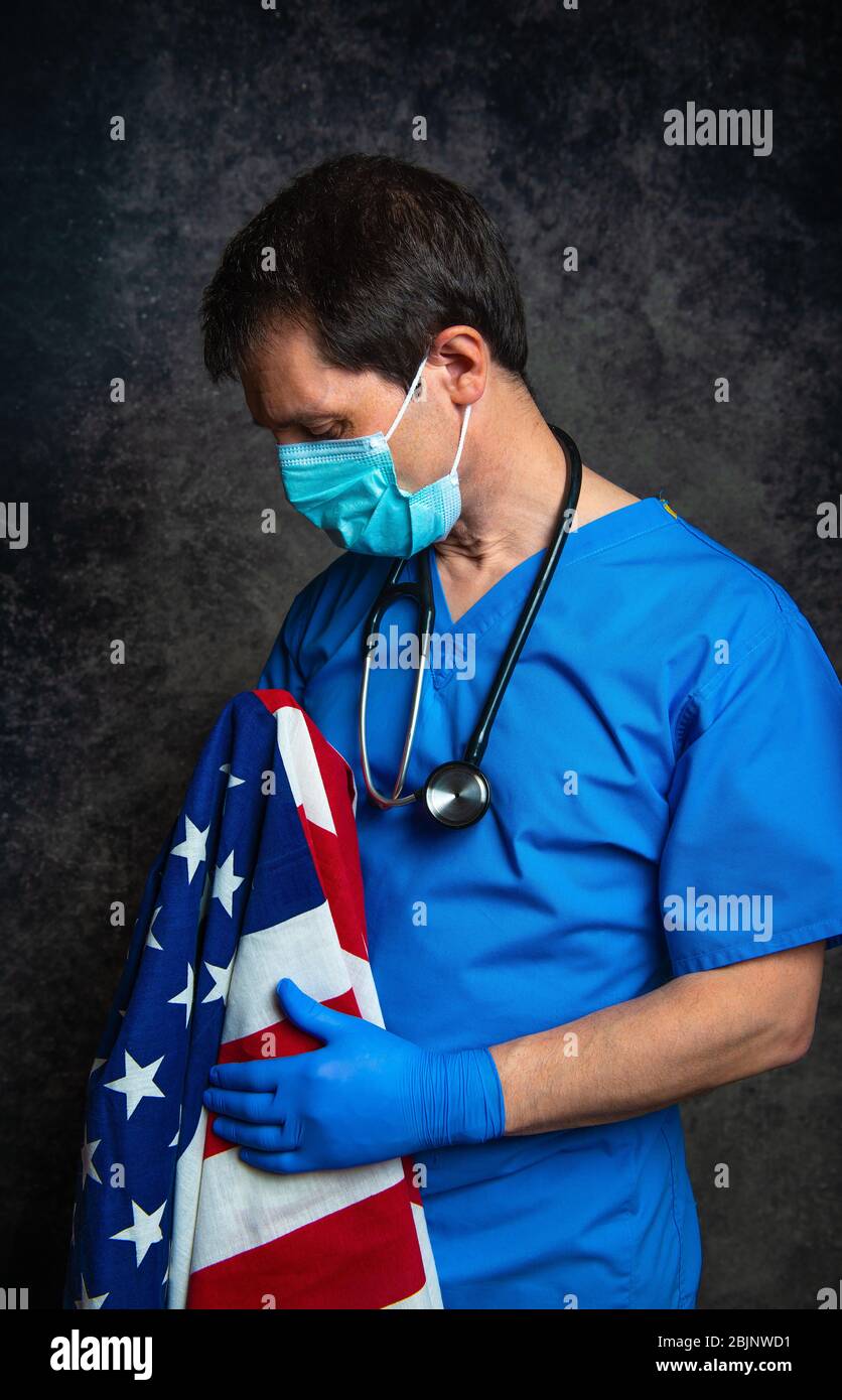 Sad/pensive male doctor in blue hospital scrubs with face mask and stethoscope, nursing the Stars & Stripes American flag close to his chest. Stock Photo