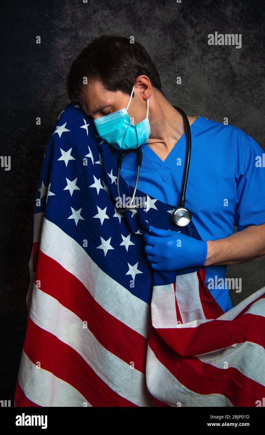 Sad/pensive male doctor in blue hospital scrubs with face mask and stethoscope, nursing the Stars & Stripes American flag close to his chest. Stock Photo