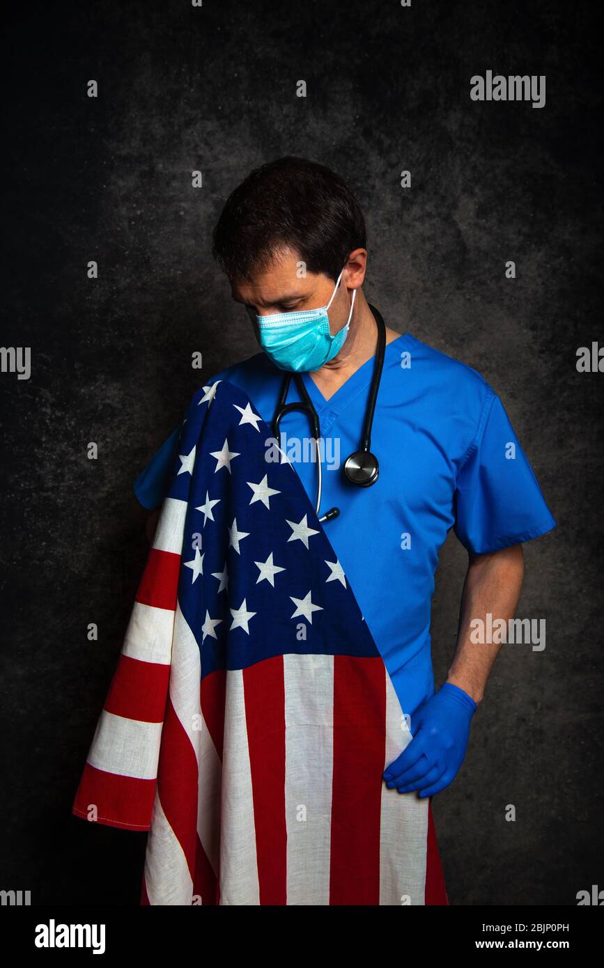 Sad/pensive male doctor in blue hospital scrubs with face mask and stethoscope, nursing the Stars & Stripes American flag close to his chest. Stock Photo