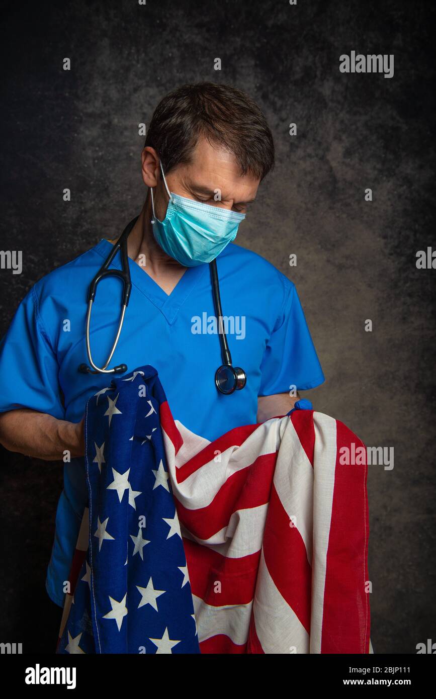 Sad/pensive male doctor in blue hospital scrubs with face mask and stethoscope, nursing the Stars & Stripes American flag close to his chest. Stock Photo