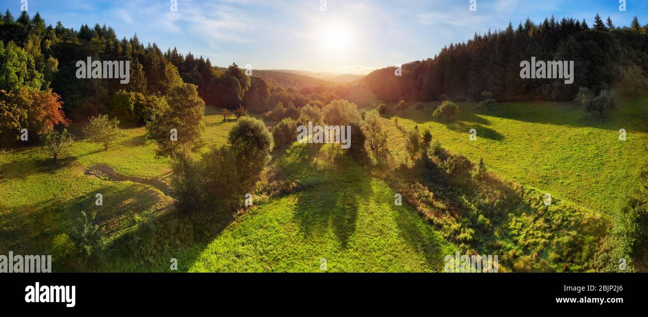 Aerial landscape panorama after sunrise: gorgeous scenery with the sun in the blue sky, trees on green meadows casting long shadows, surrounded by for Stock Photo