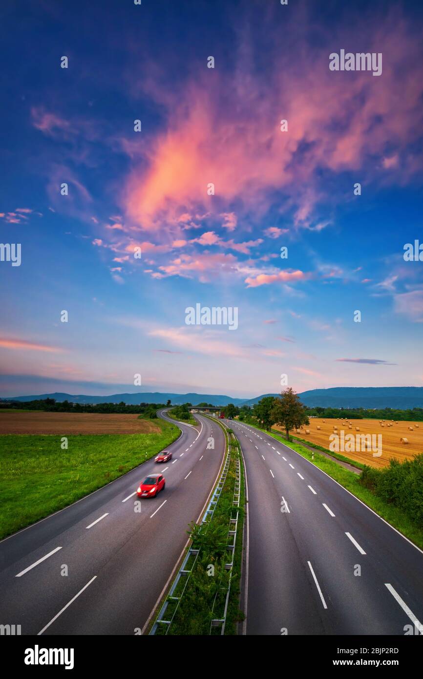 Wide, almost empty road under the colorful sky after sunset, with beautiful red clouds and two red cars with slight motion blur, a dynamic transportat Stock Photo
