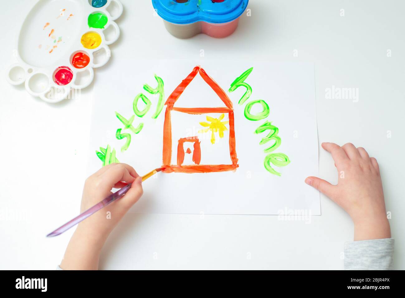 Top view child is drawing red house by watercolors with words Stay Home on white sheet of paper. Stay Home concept. Stock Photo