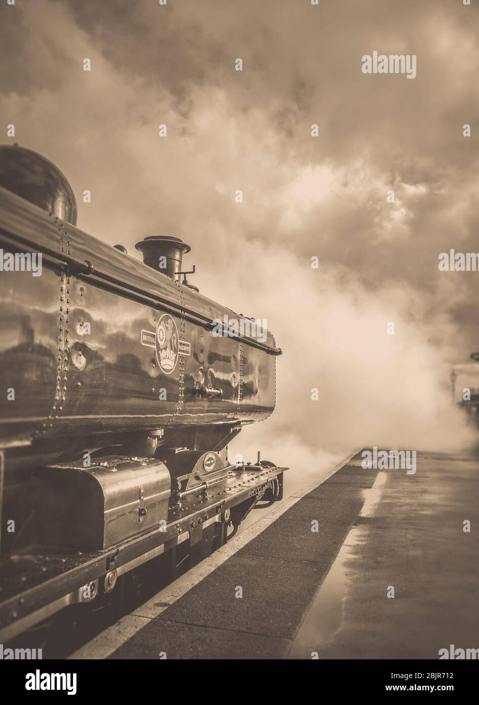 Sepia side view of vintage UK steam train near platform, leaving heritage railway station. Fulll steam ahead for this atmospheric departure photo! Stock Photo