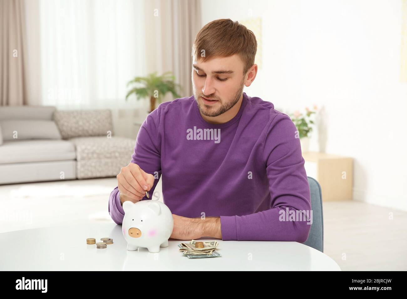 Man putting money into piggy bank indoors Stock Photo - Alamy
