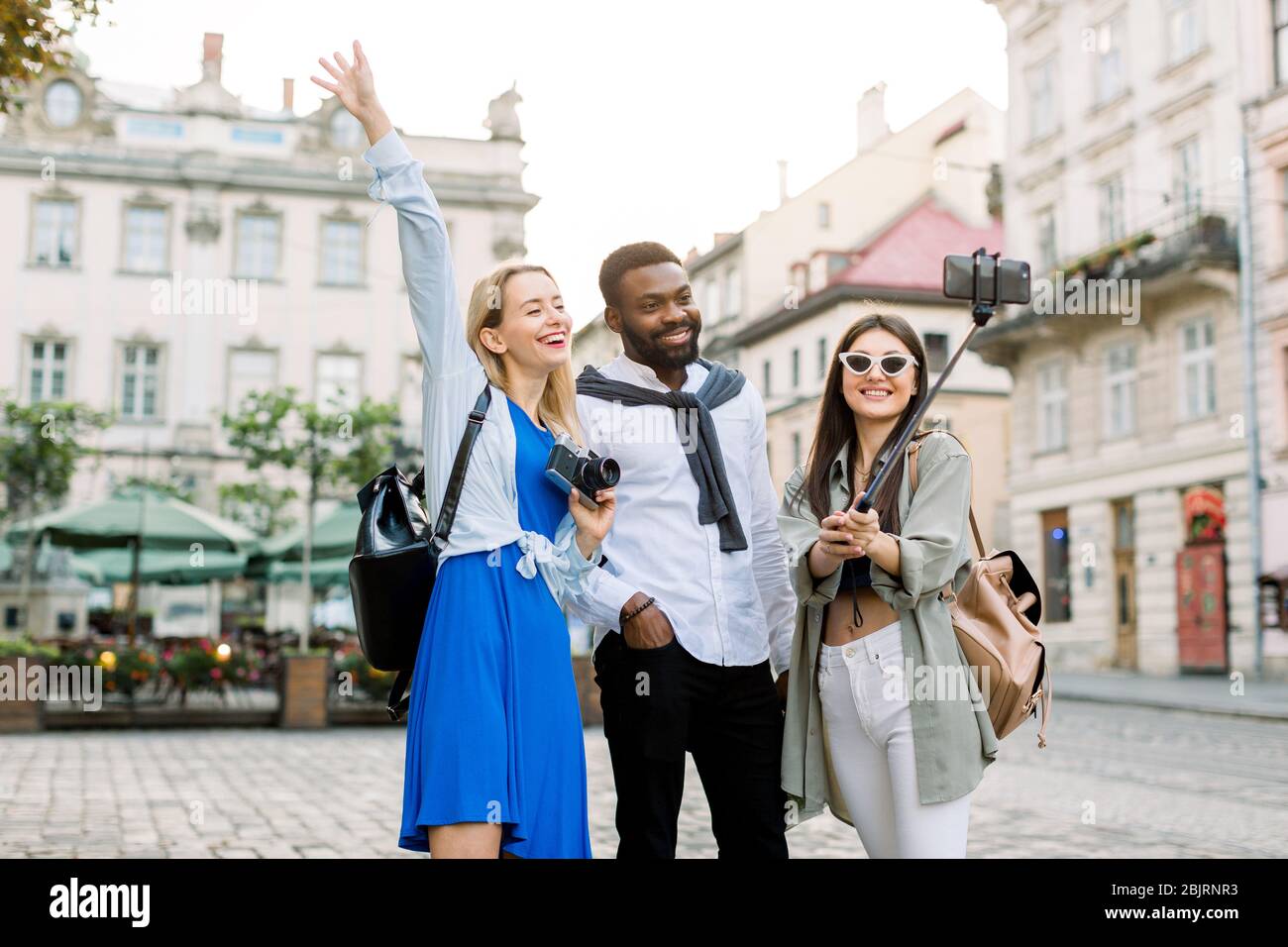 Excited cheerful mix raced friends enjoying walking outdoors in European city. Man and two women walking outside, hugging each other, laughing and Stock Photo