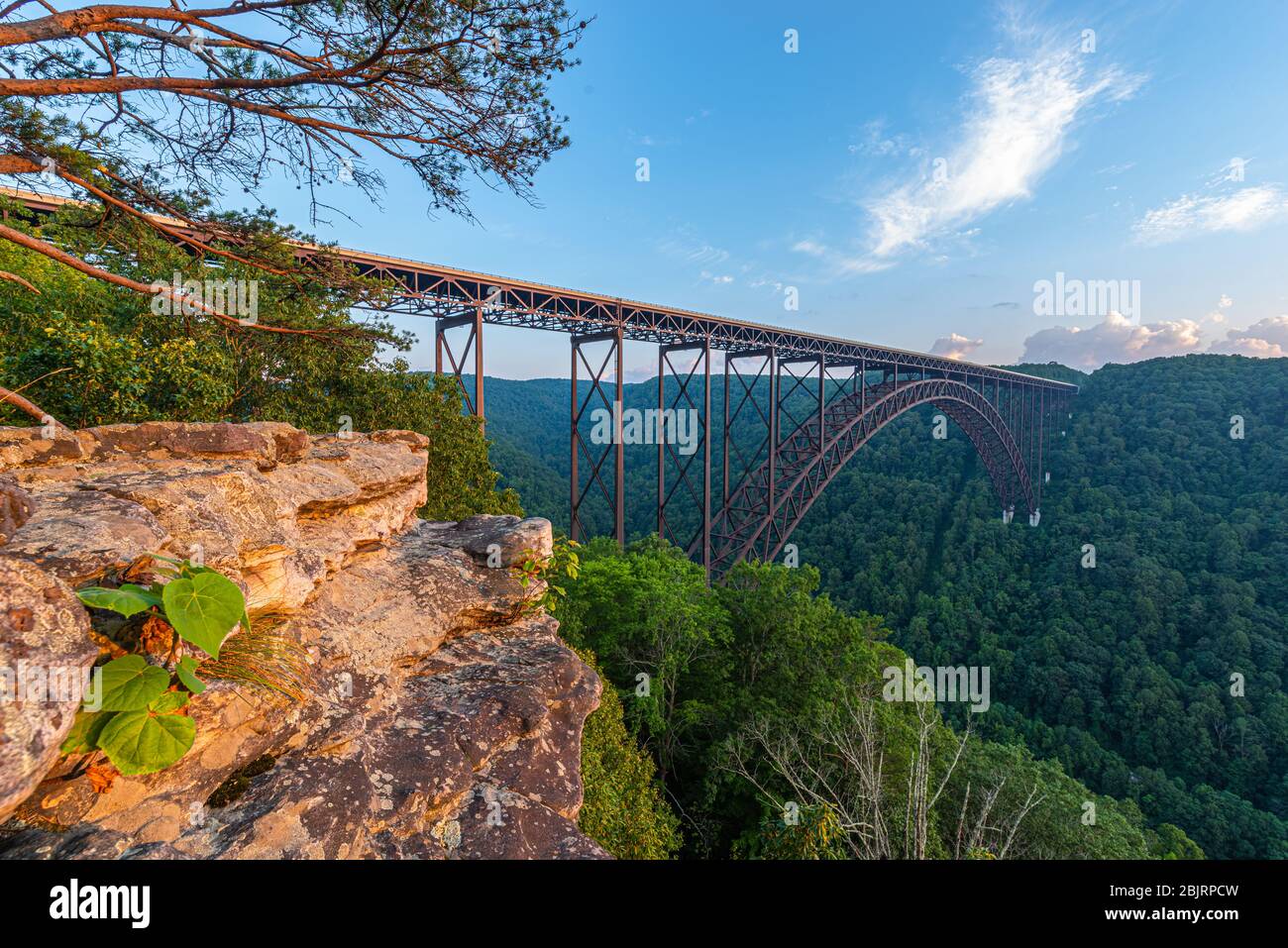 The large steel arch bridge of the New River Gorge spans the canyon below a blue sky viewed from the rock ledge of a climbing area. Stock Photo