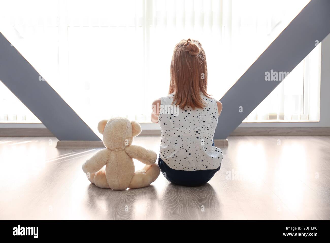 Little girl with teddy bear sitting on floor near window in empty room ...