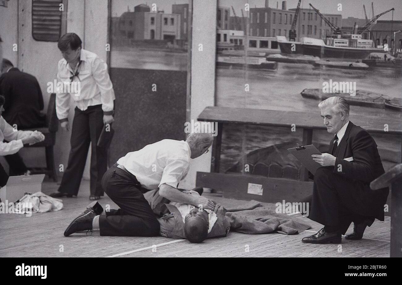 1980s, historical, Inside a hall, people at a first aid course being marked by examiners of officials on the practical elements, with a male first aider in the picture giving chest compressions or CPR to a man lying on the floor, England, UK. Stock Photo