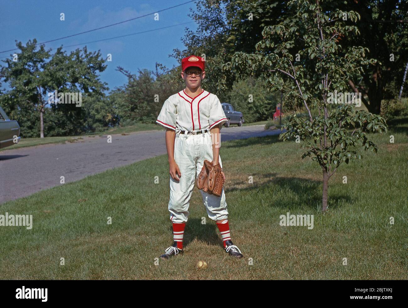 A young boy poses in his front garden wearing his baseball kit, USA c. 1960. He has his baseball mitt on, his white uniform has red trim and he a red cap and wears red socks. A baseball is at his feet. Players on a baseball team wear matching uniforms. Originally a team was identified by the colours of their stockings and the success of the Cincinnati Red Stockings popularised the adoption of sock colour as the club identity. Stock Photo