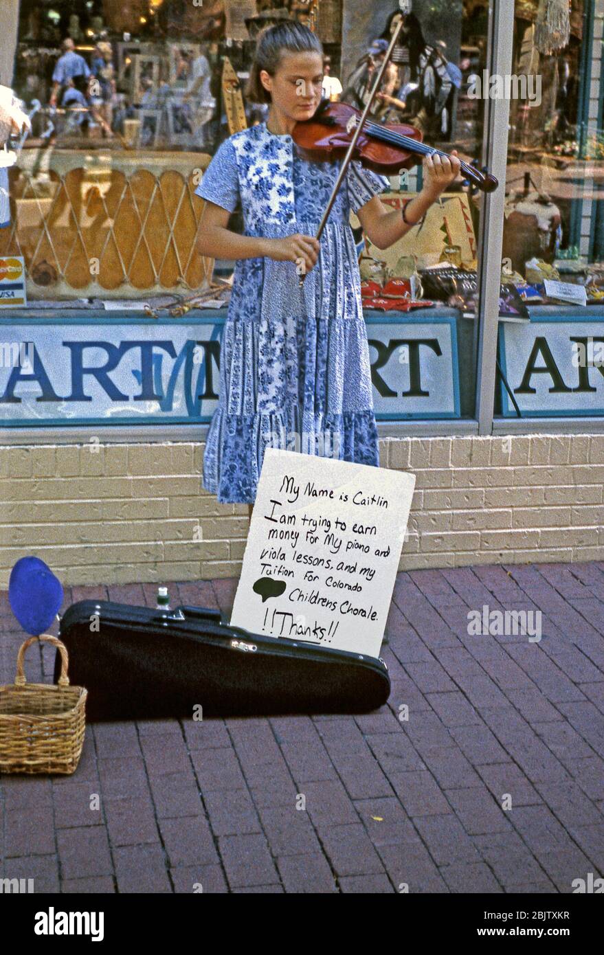 A street musician busking in Denver, Colorado, USA c. 1965. She is playing a violin and a hand-written sign at her feet tells us that she is called Caitlin and is busking to collect money to pay for piano and viola lessons plus tuition for the Colorado Childrens' Chorale. She has also drawn a big heart on it. Stock Photo