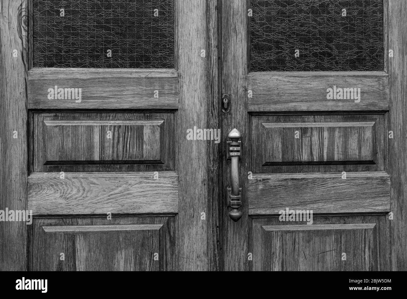 Black and white photo of wooden double door panels with rectangular windows and matte patterned glass inside frames. Retro door knob and keyhole of ol Stock Photo