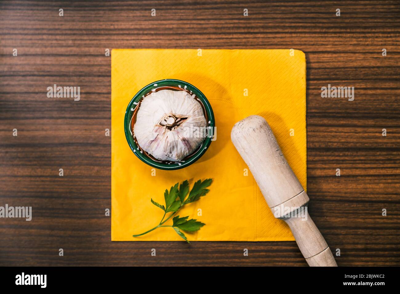 Garlic head and a parsley leaf together with a hand of mortar on top in a traditional style wooden countertop. Preparing a recipe with garlic to make Stock Photo