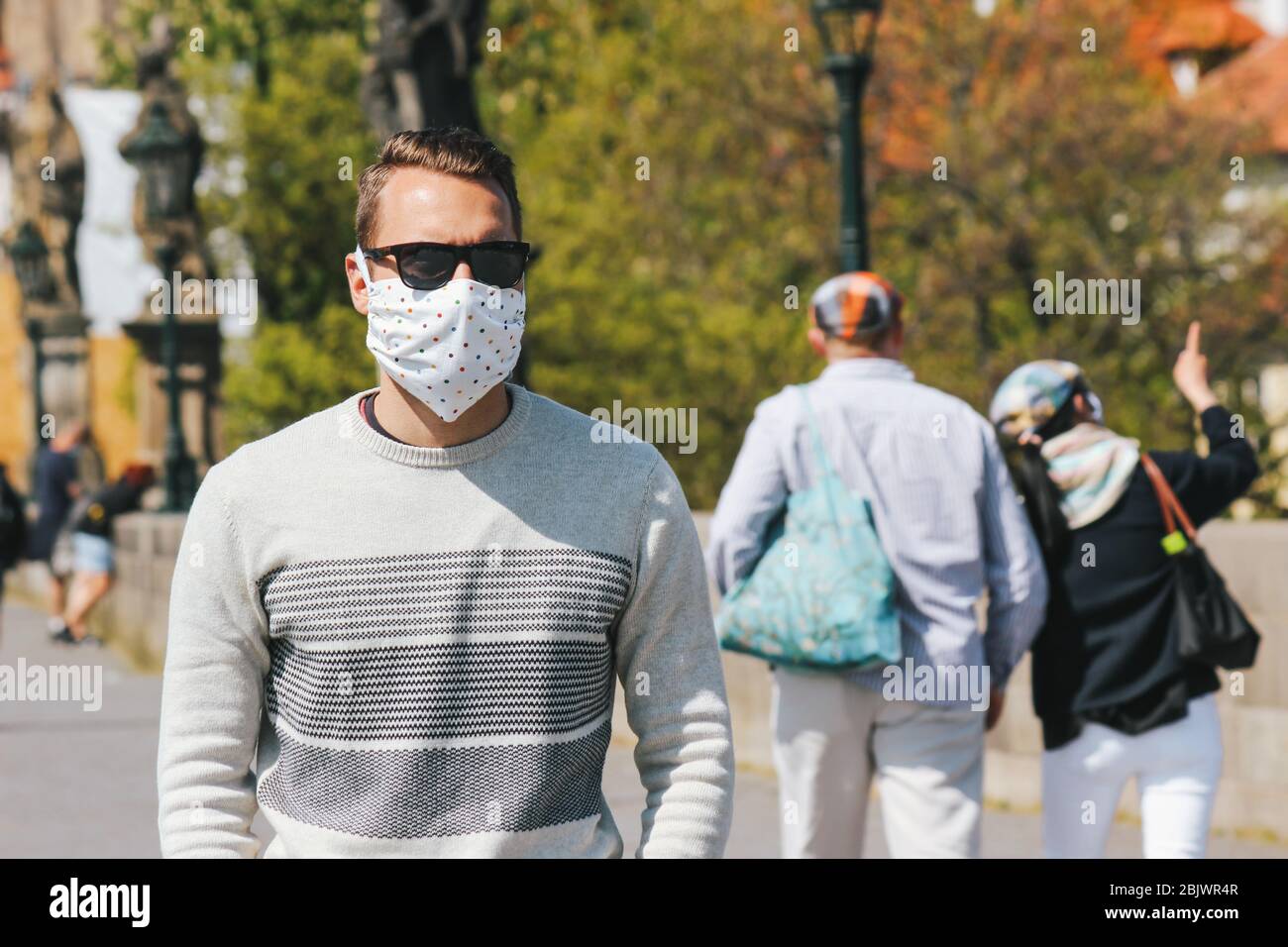 Young man with sunglasses and sewed fabric face mask photographed on the Charles Bridge in Prague, Czech Republic. Blurred people in the background. Travelling, tourism during coronavirus. COVID-19. Stock Photo
