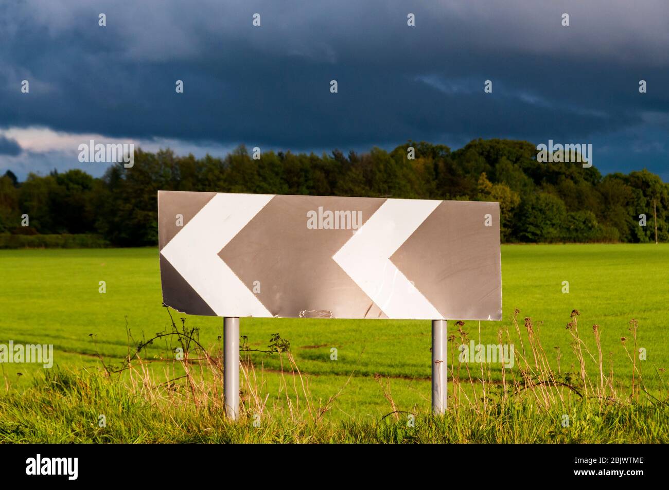 A chevron roadsign warns of a sharp bend on a country road, with dark storm clouds behind. Stock Photo