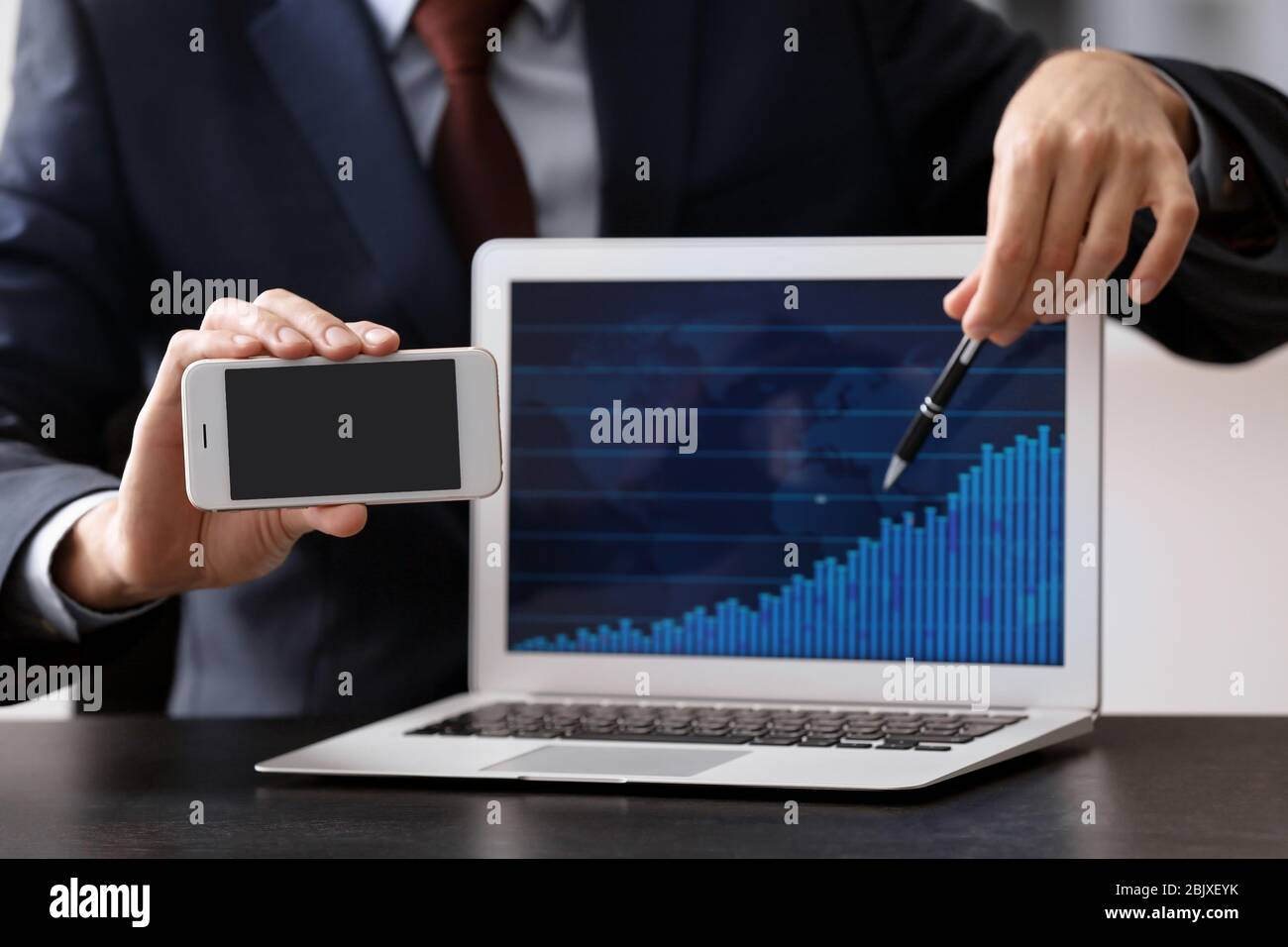 Man holding smartphone near laptop with stock chart data on screen ...