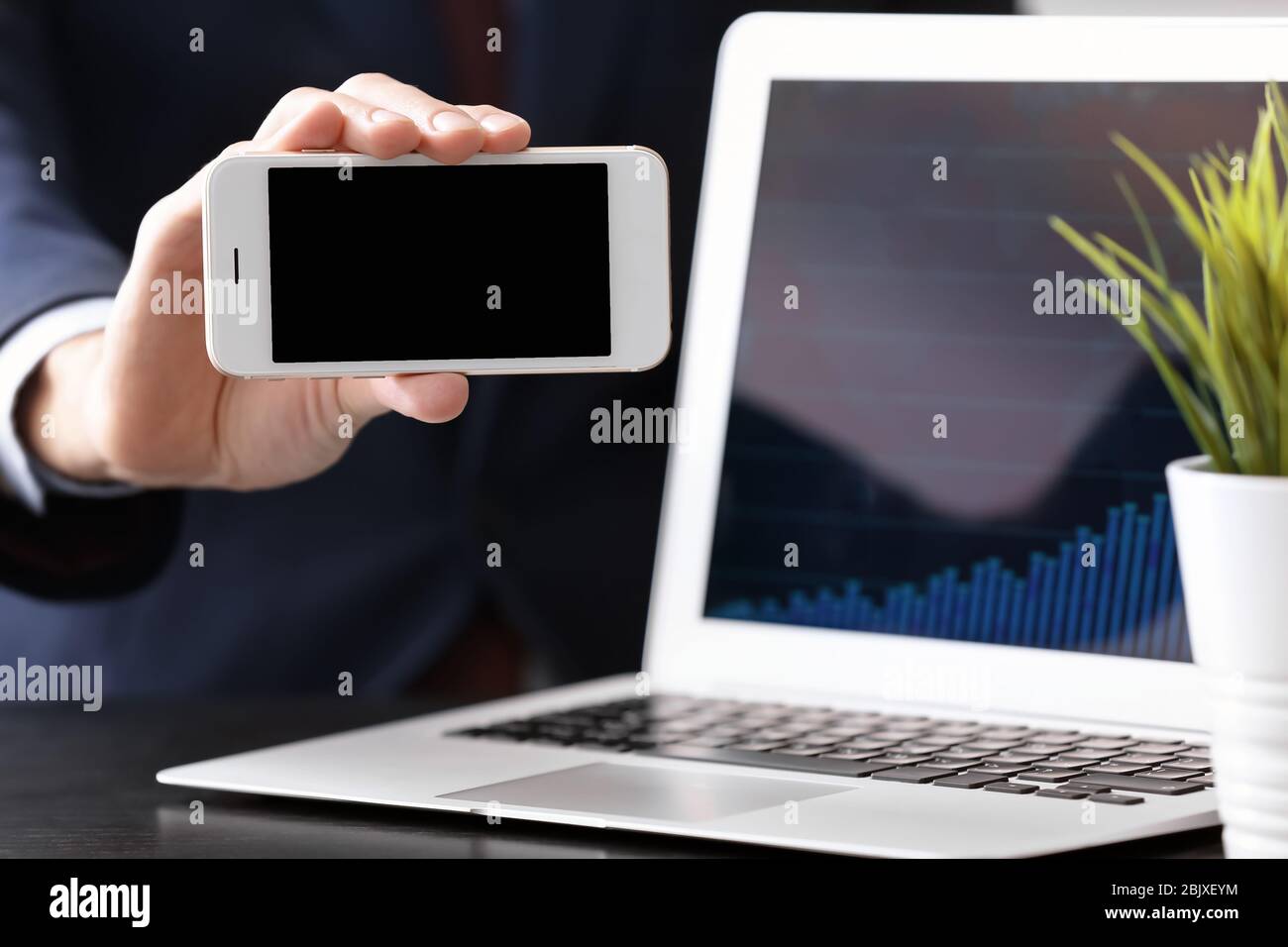 Man holding smartphone near laptop with stock chart data on screen ...