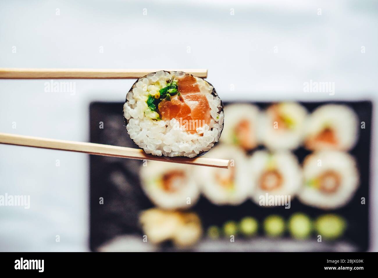 Closeup of chopsticks holding sushi infront of more sushi on a black plate over white background Stock Photo