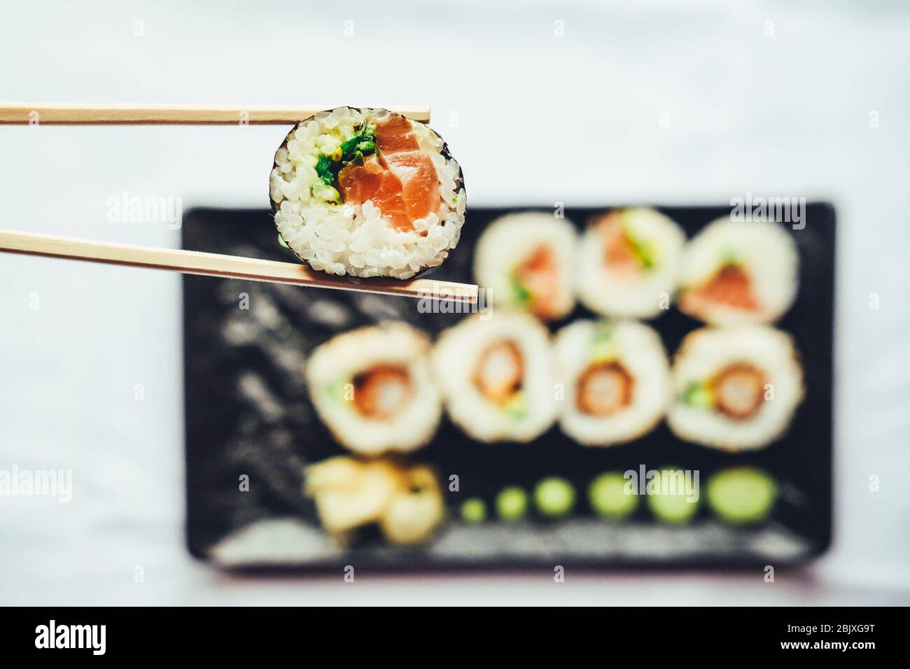 Closeup of chopsticks holding sushi infront of more sushi on a black plate over white background Stock Photo