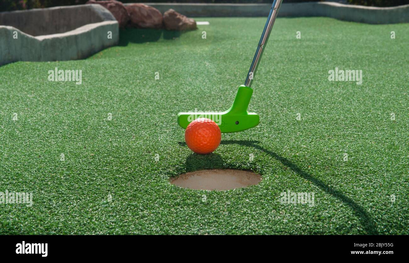 Green mini golf putter ready to tap an orange ball into the hole on green turf Stock Photo